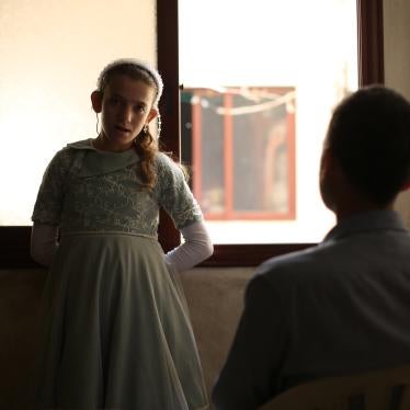 A girl stands in front of a window, looking at the camera